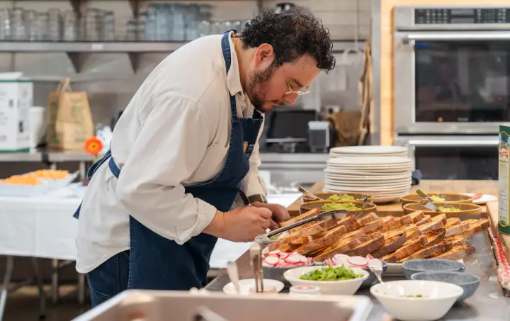 A chef writing beside bread, vegetables and prepared food on a kitchen counter.