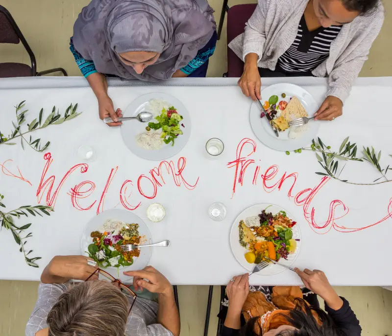 People sharing a meal around a table with a handwritten welcome.