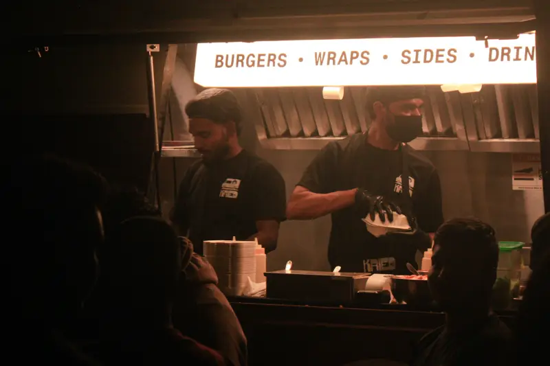 Two food-truck workers preparing and serving food through an open window at night.