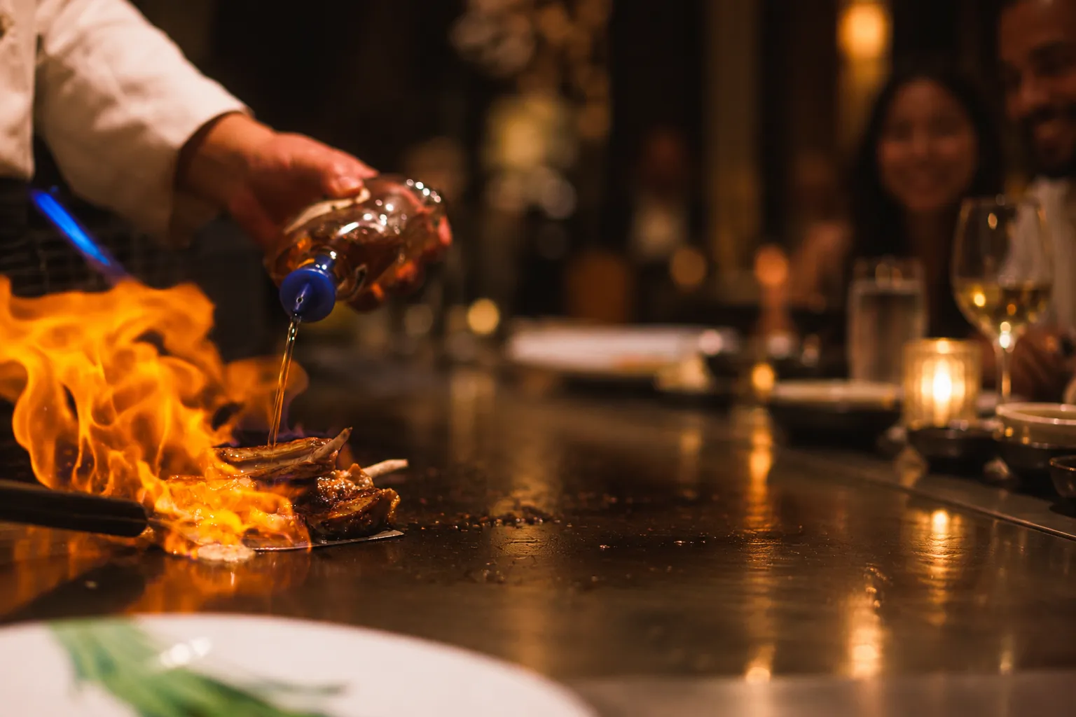 A chef finishes food over a bright teppanyaki flame, with guests softly visible beyond the grill.